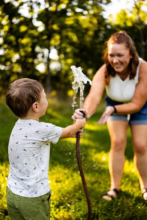 Mom and young son laughing and playing together outdoors during summertime mini session, splashing in water, chasing bubbles, and making unforgettable memories.
