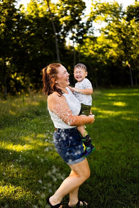Mom and young son laughing and playing together outdoors during summertime mini session, splashing in water, chasing bubbles, and making unforgettable memories.