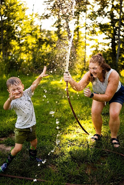 Mom and young son laughing and playing together outdoors during summertime mini session, splashing in water, chasing bubbles, and making unforgettable memories.