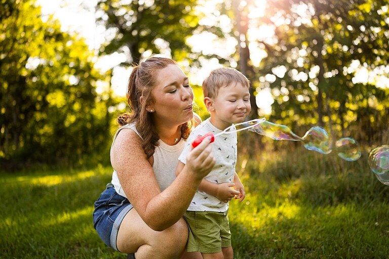 Mom and young son laughing and playing together outdoors during summertime mini session, splashing in water, chasing bubbles, and making unforgettable memories.