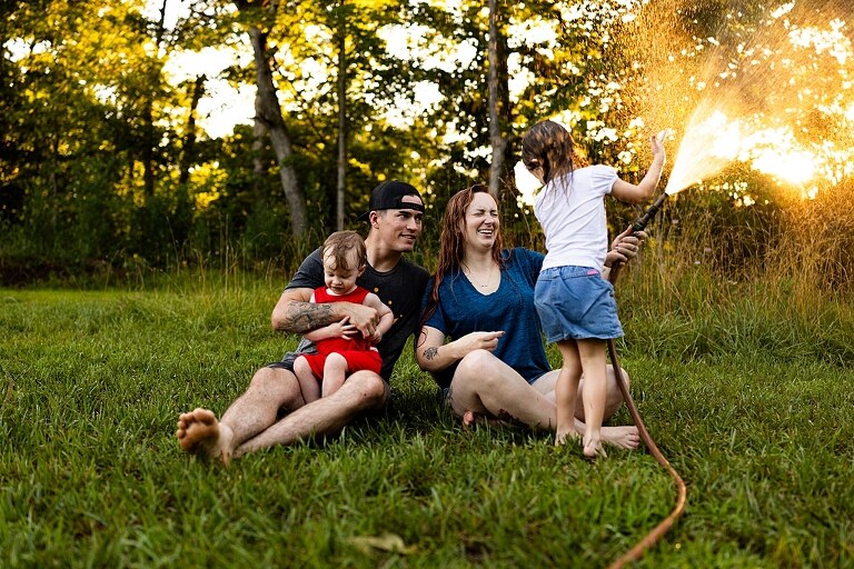 Family of four playing outdoors during summertime mini session, laughing, splashing water, blowing bubbles, and making joyful memories together.