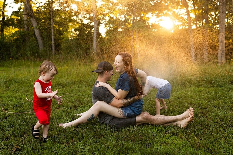 Family of four playing outdoors during summertime mini session, laughing, splashing water, blowing bubbles, and making joyful memories together.