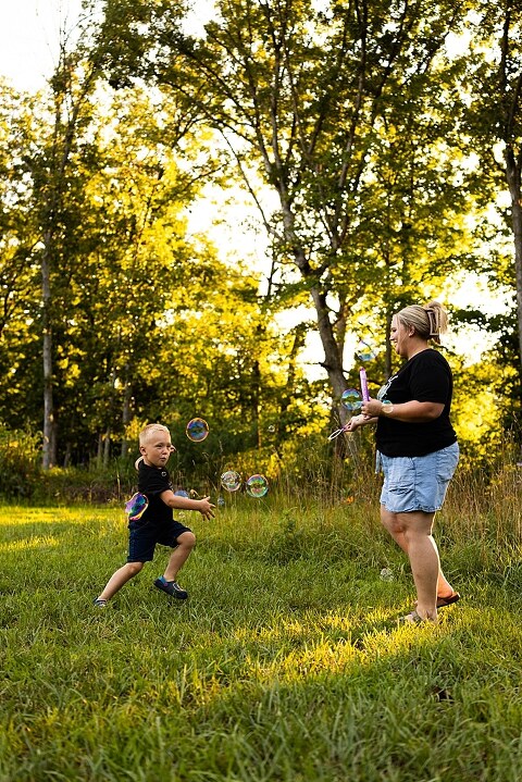 Mom and young son playing together outdoors during summertime mini session, blowing bubbles, splashing water, and sharing joyful moments in the sunshine.