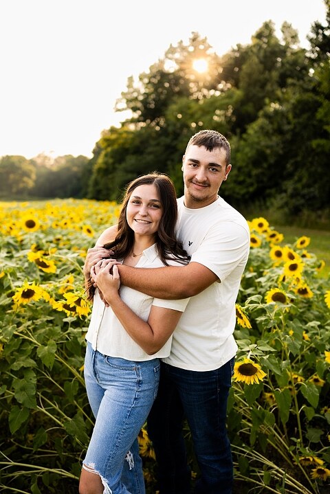 Extended family standing together in a sunflower field smiling during a summer mini session | Grandparents surrounded by their grandchildren in a bright sunflower patch | Large family group walking through sunflowers during a summer photo session