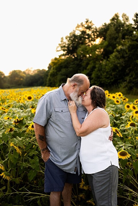 Extended family standing together in a sunflower field smiling during a summer mini session | Grandparents surrounded by their grandchildren in a bright sunflower patch | Large family group walking through sunflowers during a summer photo session