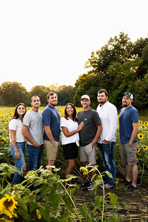Extended family standing together in a sunflower field smiling during a summer mini session | Grandparents surrounded by their grandchildren in a bright sunflower patch | Large family group walking through sunflowers during a summer photo session