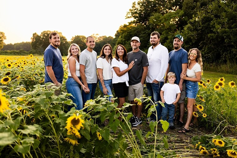Extended family standing together in a sunflower field smiling during a summer mini session | Grandparents surrounded by their grandchildren in a bright sunflower patch | Large family group walking through sunflowers during a summer photo session