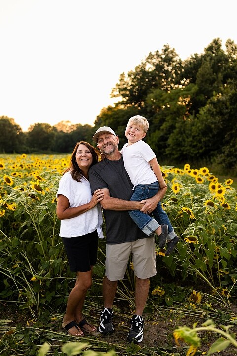 Extended family standing together in a sunflower field smiling during a summer mini session | Grandparents surrounded by their grandchildren in a bright sunflower patch | Large family group walking through sunflowers during a summer photo session