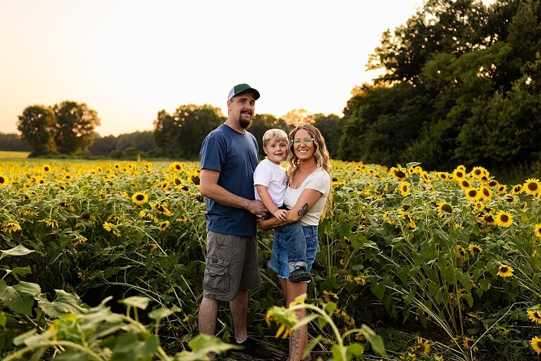 Extended family standing together in a sunflower field smiling during a summer mini session | Grandparents surrounded by their grandchildren in a bright sunflower patch | Large family group walking through sunflowers during a summer photo session