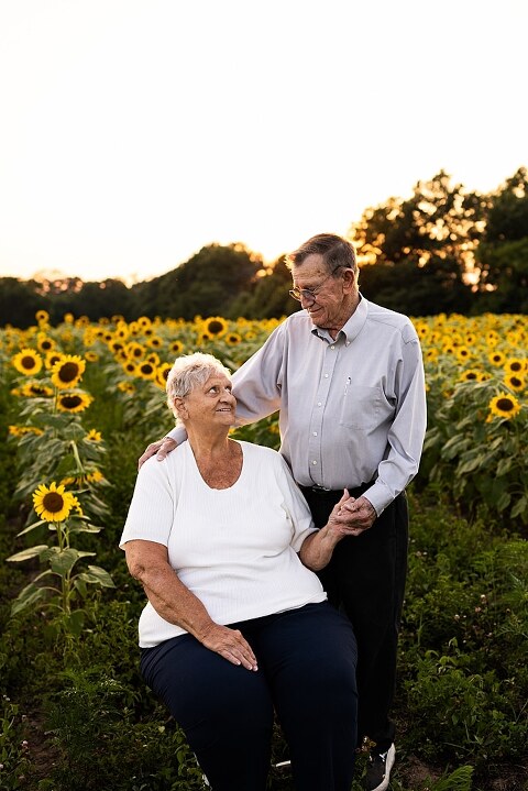 Extended family standing together in a sunflower field smiling during a summer mini session | Grandparents surrounded by their grandchildren in a bright sunflower patch | Large family group walking through sunflowers during a summer photo session