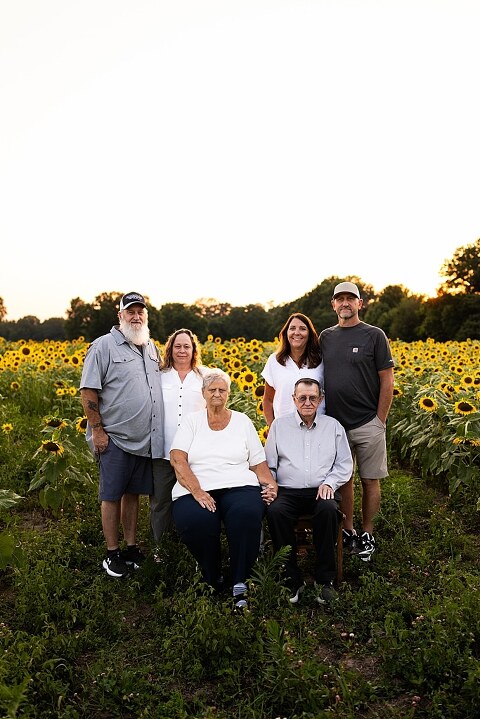 Extended family standing together in a sunflower field smiling during a summer mini session | Grandparents surrounded by their grandchildren in a bright sunflower patch | Large family group walking through sunflowers during a summer photo session