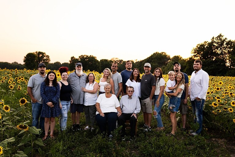 Extended family standing together in a sunflower field smiling during a summer mini session | Grandparents surrounded by their grandchildren in a bright sunflower patch | Large family group walking through sunflowers during a summer photo session
