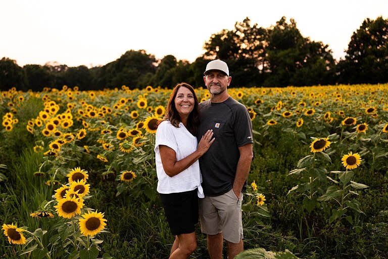 Extended family standing together in a sunflower field smiling during a summer mini session | Grandparents surrounded by their grandchildren in a bright sunflower patch | Large family group walking through sunflowers during a summer photo session