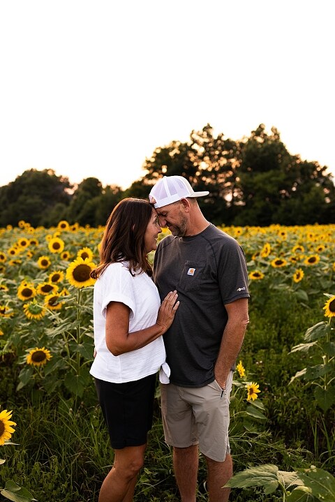 Extended family standing together in a sunflower field smiling during a summer mini session | Grandparents surrounded by their grandchildren in a bright sunflower patch | Large family group walking through sunflowers during a summer photo session
