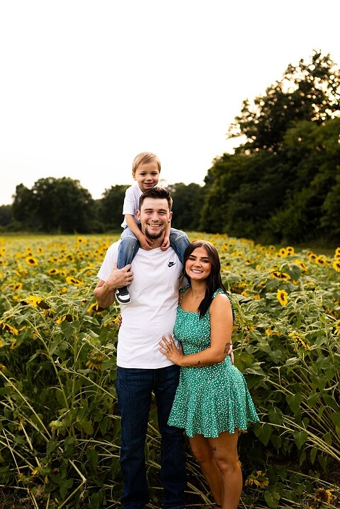 Family of three smiling together in a bright sunflower field during summer | Little boy grinning while standing among tall sunflowers
 | Parents holding their son and laughing in golden evening light | Family hugging closely surrounded by blooming sunflowers