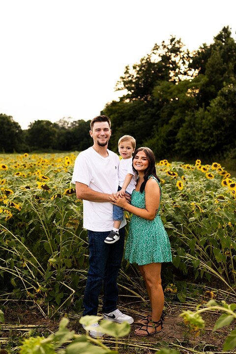 Family of three smiling together in a bright sunflower field during summer | Little boy grinning while standing among tall sunflowers
 | Parents holding their son and laughing in golden evening light | Family hugging closely surrounded by blooming sunflowers