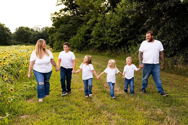 Blended family smiling together in a sunflower field during a summer mini session | Mom and children laughing among tall sunflowers in warm evening light | Parents holding hands and smiling in a bright sunflower patch | Close up of family hugging with golden blooms surrounding them