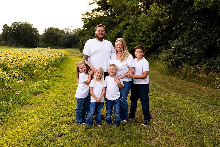 Blended family smiling together in a sunflower field during a summer mini session | Mom and children laughing among tall sunflowers in warm evening light | Parents holding hands and smiling in a bright sunflower patch | Close up of family hugging with golden blooms surrounding them
