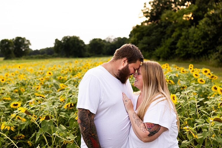 Blended family smiling together in a sunflower field during a summer mini session | Mom and children laughing among tall sunflowers in warm evening light | Parents holding hands and smiling in a bright sunflower patch | Close up of family hugging with golden blooms surrounding them
