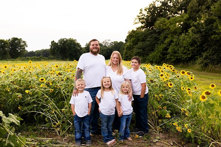 Blended family smiling together in a sunflower field during a summer mini session | Mom and children laughing among tall sunflowers in warm evening light | Parents holding hands and smiling in a bright sunflower patch | Close up of family hugging with golden blooms surrounding them