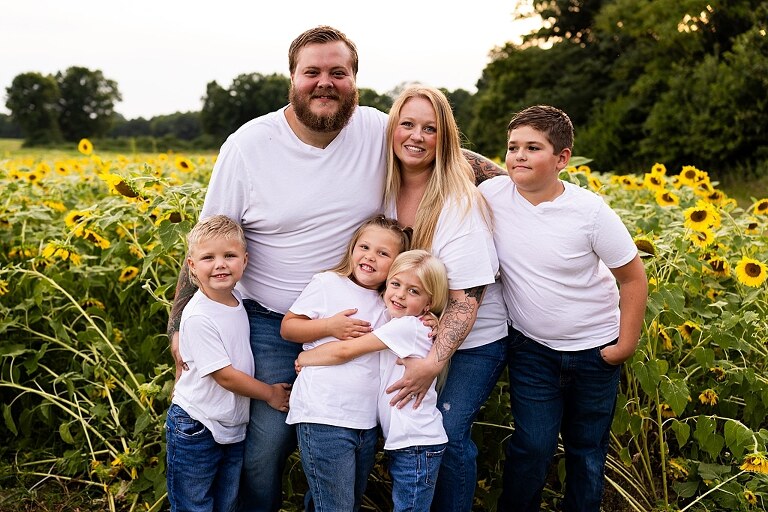 Blended family smiling together in a sunflower field during a summer mini session | Mom and children laughing among tall sunflowers in warm evening light | Parents holding hands and smiling in a bright sunflower patch | Close up of family hugging with golden blooms surrounding them