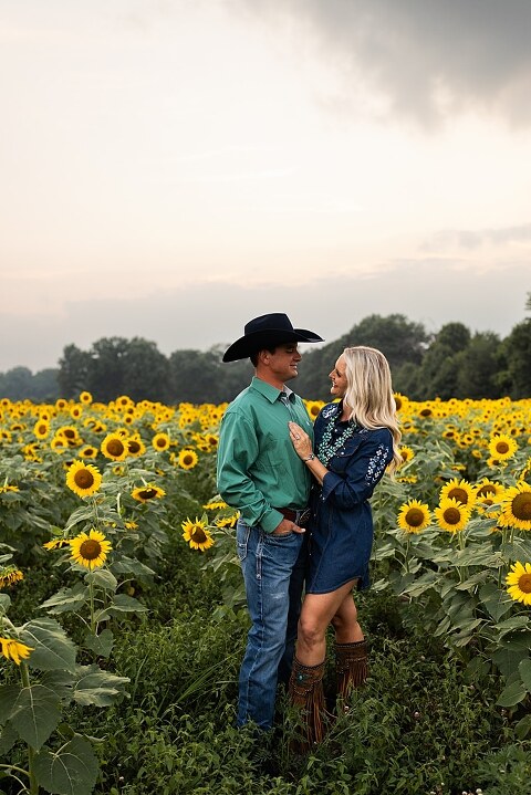 Couple standing together in a sunflower field during sunset in Jonesburg Missouri | Dog sitting in front of sunflowers with golden evening light | Couple laughing while holding their dog in a sunflower field | Sunflower Mini Photo Shoot with couple and dog at golden hour | Dog running through sunflower rows during family photo session | Missouri Sunflower Photographer