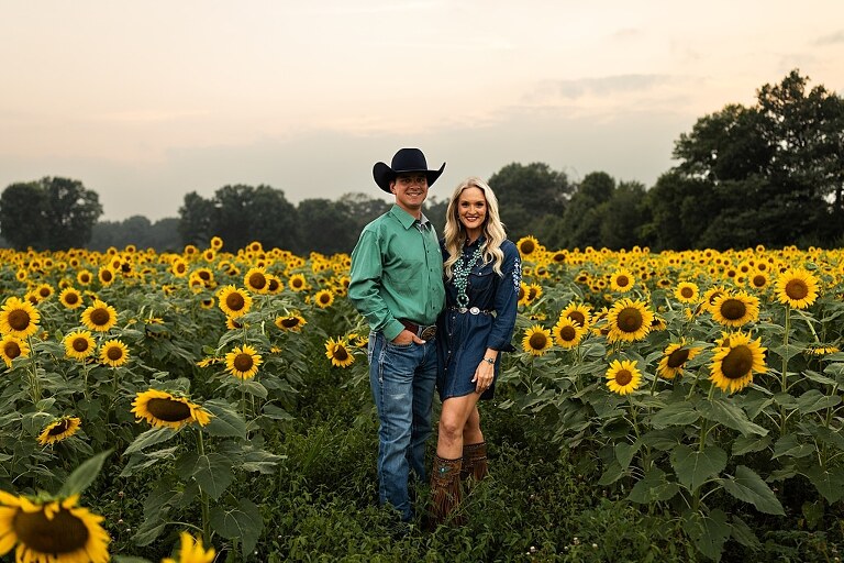 Couple standing together in a sunflower field during sunset in Jonesburg Missouri | Dog sitting in front of sunflowers with golden evening light | Couple laughing while holding their dog in a sunflower field | Sunflower Mini Photo Shoot with couple and dog at golden hour | Dog running through sunflower rows during family photo session | Missouri Sunflower Photographer