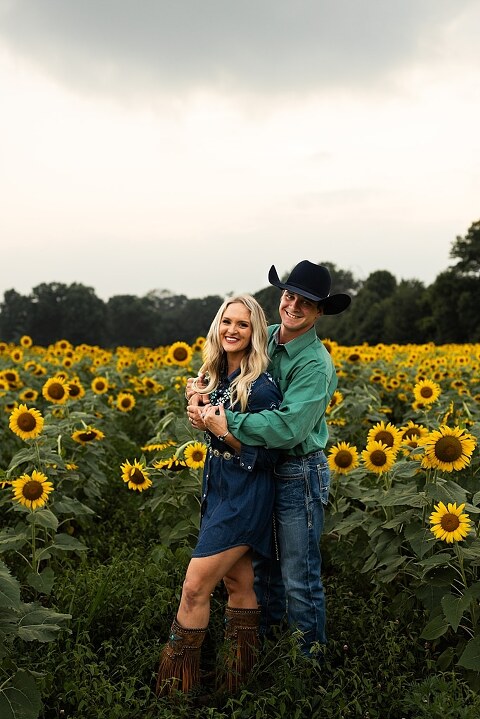 Couple standing together in a sunflower field during sunset in Jonesburg Missouri | Dog sitting in front of sunflowers with golden evening light | Couple laughing while holding their dog in a sunflower field | Sunflower Mini Photo Shoot with couple and dog at golden hour | Dog running through sunflower rows during family photo session | Missouri Sunflower Photographer
