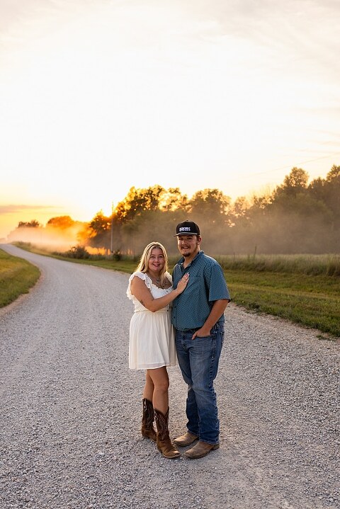 Couple walking hand in hand down a gravel road at sunset | Woman smiling at her partner during a country photo session | Former Missouri State Fair Queen posing with her partner in soft evening light | Close up of couple laughing while standing on a quiet country road
