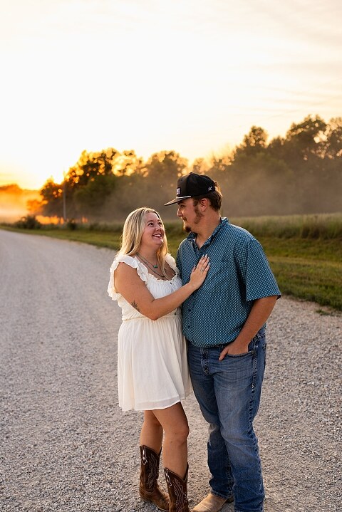 Couple walking hand in hand down a gravel road at sunset | Woman smiling at her partner during a country photo session | Former Missouri State Fair Queen posing with her partner in soft evening light | Close up of couple laughing while standing on a quiet country road