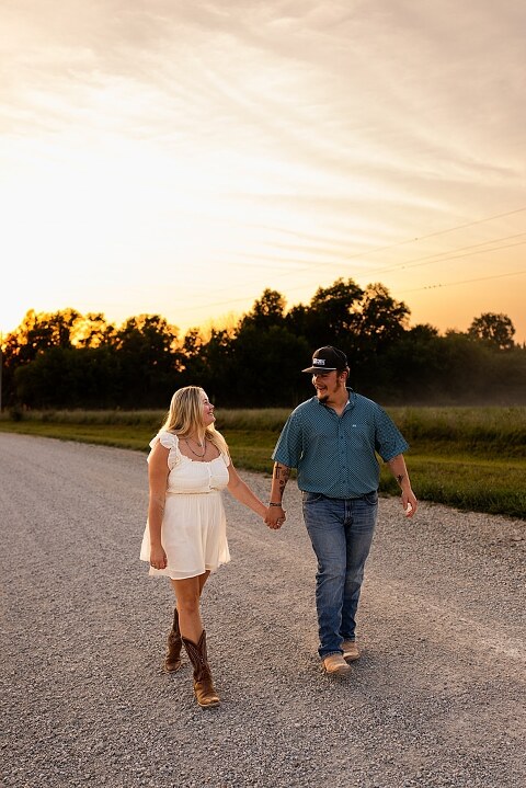 Couple walking hand in hand down a gravel road at sunset | Woman smiling at her partner during a country photo session | Former Missouri State Fair Queen posing with her partner in soft evening light | Close up of couple laughing while standing on a quiet country road