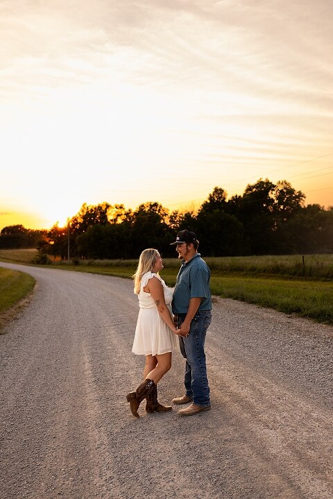 Couple walking hand in hand down a gravel road at sunset | Woman smiling at her partner during a country photo session | Former Missouri State Fair Queen posing with her partner in soft evening light | Close up of couple laughing while standing on a quiet country road