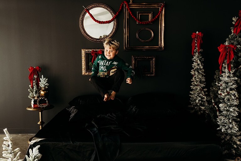 Moody Christmas mini session setup with a black bed and snow-dusted trees.
Five-year-old boy smiling on the Christmas mini session bed.
Close-up of cranberry and popcorn garland on the studio set.
Child surrounded by gold and red Christmas accents in the studio.
Full view of the 2025 Christmas Minis setup in Lake St. Louis Missouri.