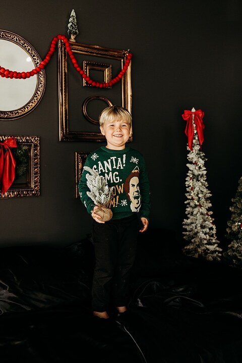 Moody Christmas mini session setup with a black bed and snow-dusted trees.
Five-year-old boy smiling on the Christmas mini session bed.
Close-up of cranberry and popcorn garland on the studio set.
Child surrounded by gold and red Christmas accents in the studio.
Full view of the 2025 Christmas Minis setup in Lake St. Louis Missouri.