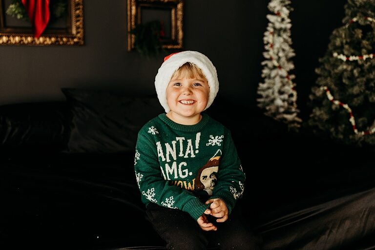 Moody Christmas mini session setup with a black bed and snow-dusted trees.
Five-year-old boy smiling on the Christmas mini session bed.
Close-up of cranberry and popcorn garland on the studio set.
Child surrounded by gold and red Christmas accents in the studio.
Full view of the 2025 Christmas Minis setup in Lake St. Louis Missouri.