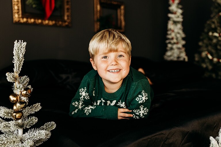 Moody Christmas mini session setup with a black bed and snow-dusted trees.
Five-year-old boy smiling on the Christmas mini session bed.
Close-up of cranberry and popcorn garland on the studio set.
Child surrounded by gold and red Christmas accents in the studio.
Full view of the 2025 Christmas Minis setup in Lake St. Louis Missouri.