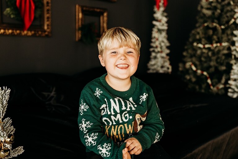 Moody Christmas mini session setup with a black bed and snow-dusted trees.
Five-year-old boy smiling on the Christmas mini session bed.
Close-up of cranberry and popcorn garland on the studio set.
Child surrounded by gold and red Christmas accents in the studio.
Full view of the 2025 Christmas Minis setup in Lake St. Louis Missouri.