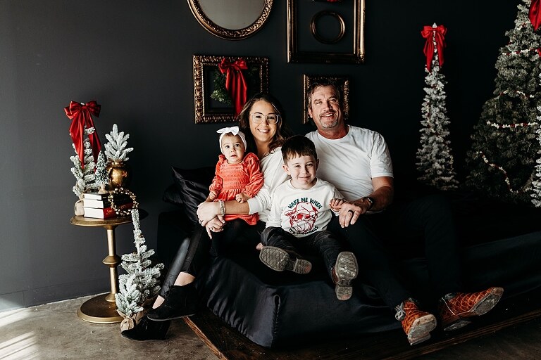 Oden family posing in a moody Christmas mini session setup with black bed, snowy trees, and nostalgic garlands in a Lake St Louis studio