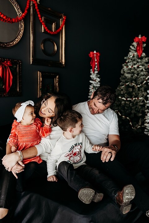 Oden family posing in a moody Christmas mini session setup with black bed, snowy trees, and nostalgic garlands in a Lake St Louis studio