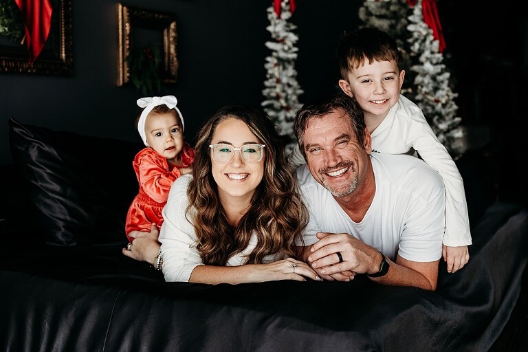 Oden family posing in a moody Christmas mini session setup with black bed, snowy trees, and nostalgic garlands in a Lake St Louis studio