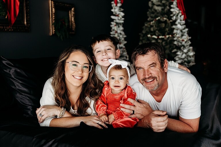 Oden family posing in a moody Christmas mini session setup with black bed, snowy trees, and nostalgic garlands in a Lake St Louis studio
