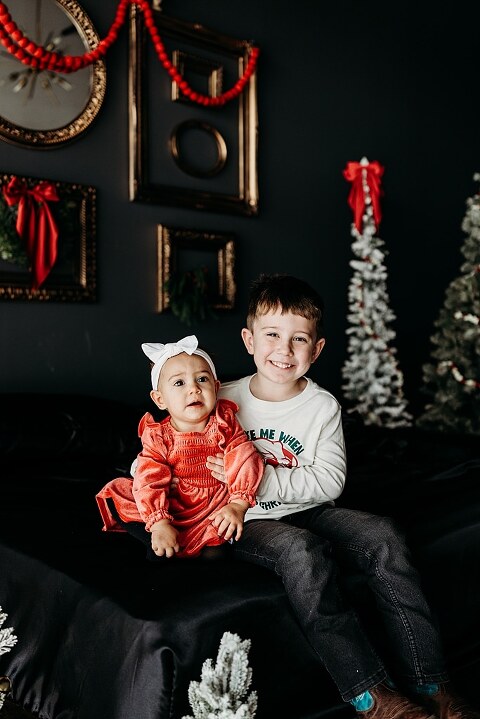 Oden family posing in a moody Christmas mini session setup with black bed, snowy trees, and nostalgic garlands in a Lake St Louis studio