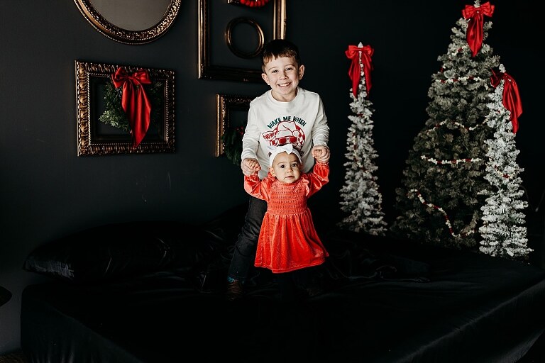 Oden family posing in a moody Christmas mini session setup with black bed, snowy trees, and nostalgic garlands in a Lake St Louis studio