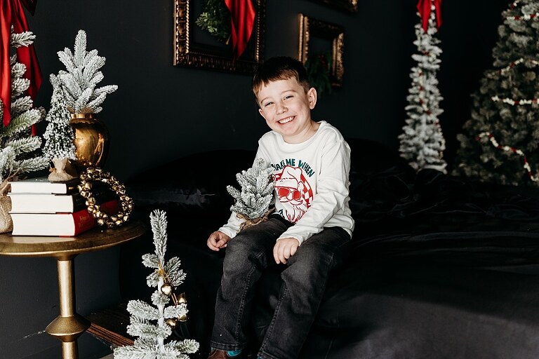 Oden family posing in a moody Christmas mini session setup with black bed, snowy trees, and nostalgic garlands in a Lake St Louis studio