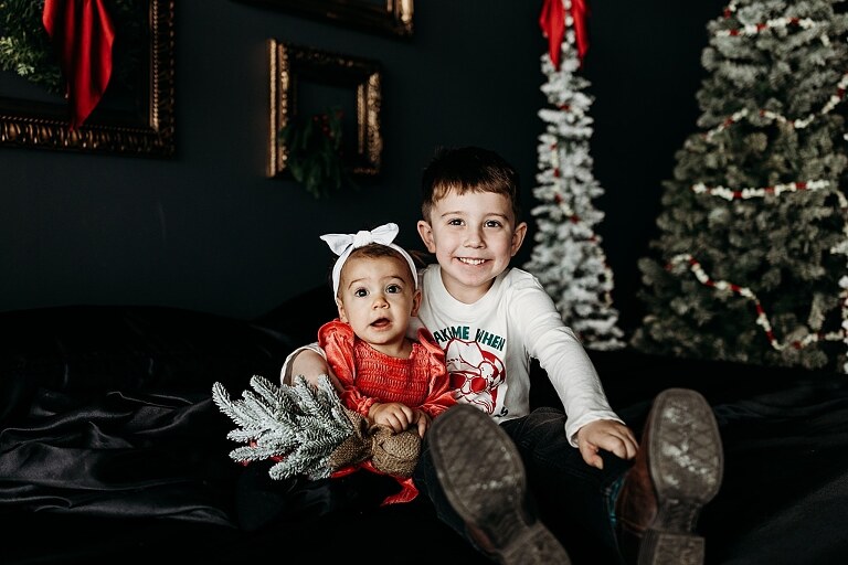 Oden family posing in a moody Christmas mini session setup with black bed, snowy trees, and nostalgic garlands in a Lake St Louis studio