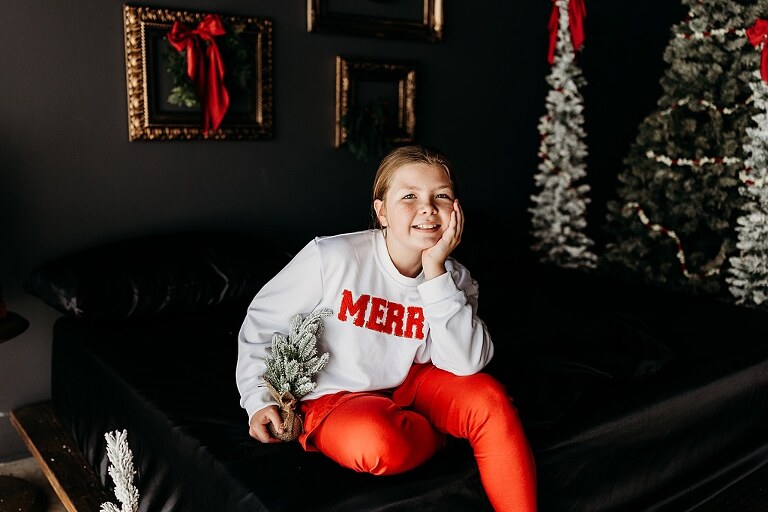 Moody Christmas mini session setup with black bed, snowy Christmas trees, cranberry and popcorn garland, and gold and red holiday accents in a Lake St Louis studio