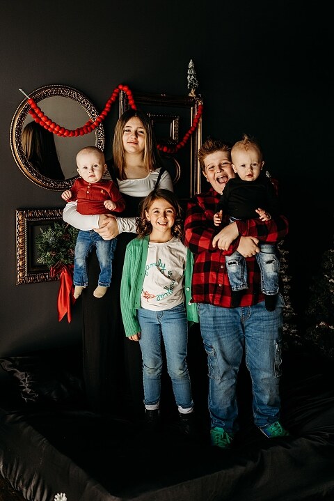 Family sitting on black bed during Christmas Mini Session with trees and garland
Parents holding newborn twins in studio with red and gold holiday details
Children sitting together on bed with Christmas trees behind them
Close up of popcorn and cranberry garland in Christmas Mini setup