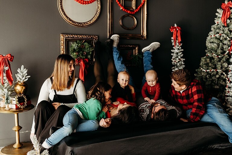 Family sitting on black bed during Christmas Mini Session with trees and garland
Parents holding newborn twins in studio with red and gold holiday details
Children sitting together on bed with Christmas trees behind them
Close up of popcorn and cranberry garland in Christmas Mini setup